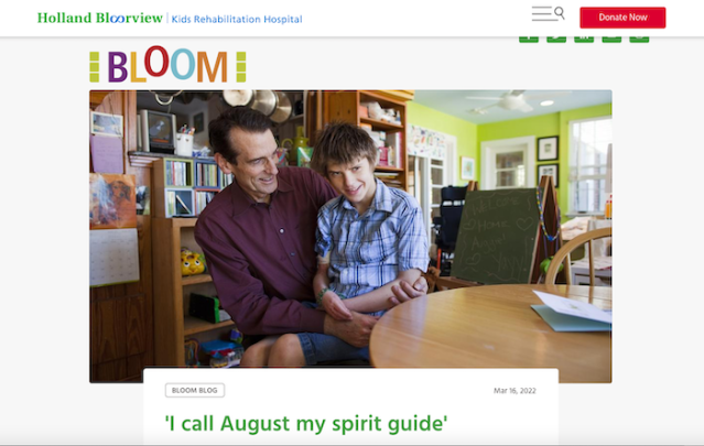 Gabbard holding his son on his lap. Both are smiling. They are in a cluttered room in their house on a sunny day.